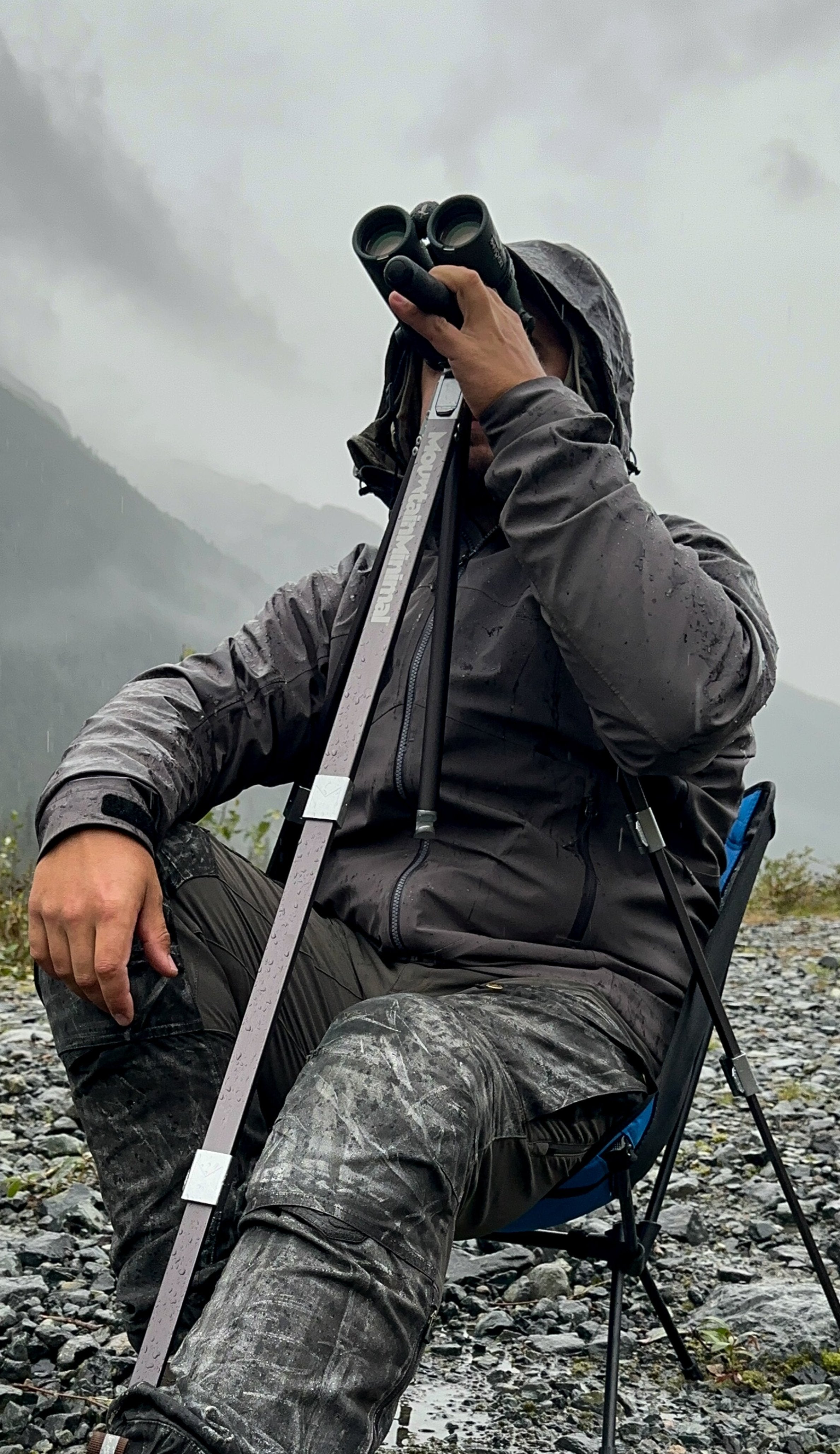 Mountain goat guide sits in rain and glasses through binoculars from a Mountain Minimal backcountry hunting tripod.